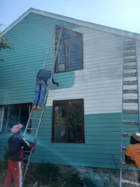 Painters on ladders applying teal paint to a house exterior